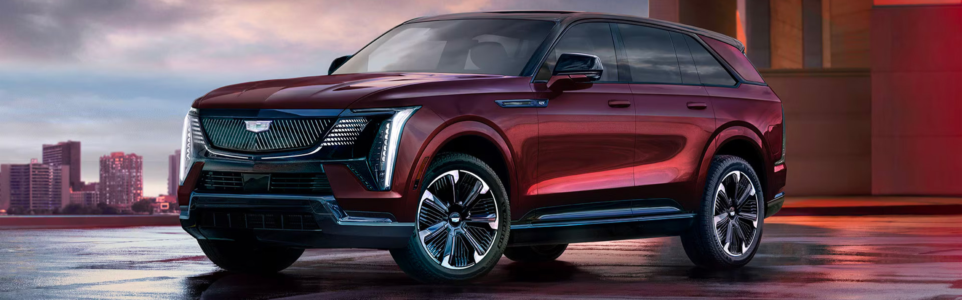 2025 Cadillac Escalade IQ electric SUV shown in a deep red finish, parked on a wet urban plaza at dusk.