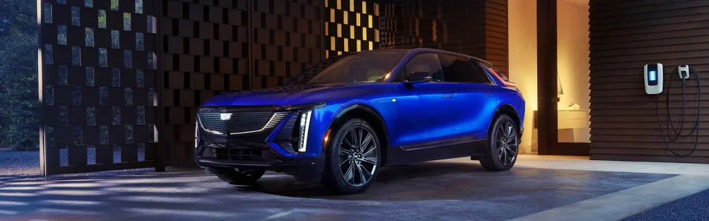 This image shows the 2026 Cadillac Lyriq in a bold blue color, parked in a modern driveway with a charging station beside it, highlighting its futuristic design.