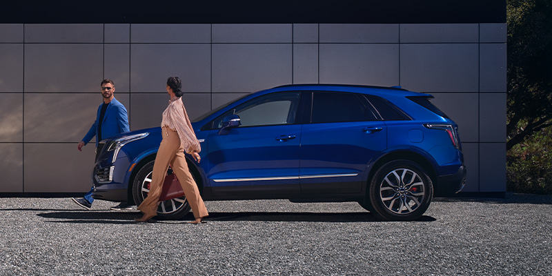 2026 Cadillac XT6 SUV parked beside a modern building in West Amarillo, TX, with a stylish couple walking past, highlighting luxury design and upscale appeal.