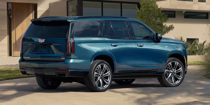 Blue Cadillac Escalade parked in a driveway in front of a modern house. The car has sleek lines and shiny alloy wheels, conveying a sense of luxury and style.